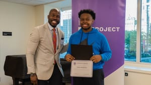 Conyers poses with a student, who is holding a diploma, for a snapshot in an indoor space, a piano behind them