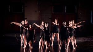 A dance ensemble of nine women in short black costumes pose together, facing front with one arm outstretched.