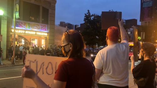 A view from behind the sign-waving protestors, with the crowded, lit-up theater opening visible across the street.