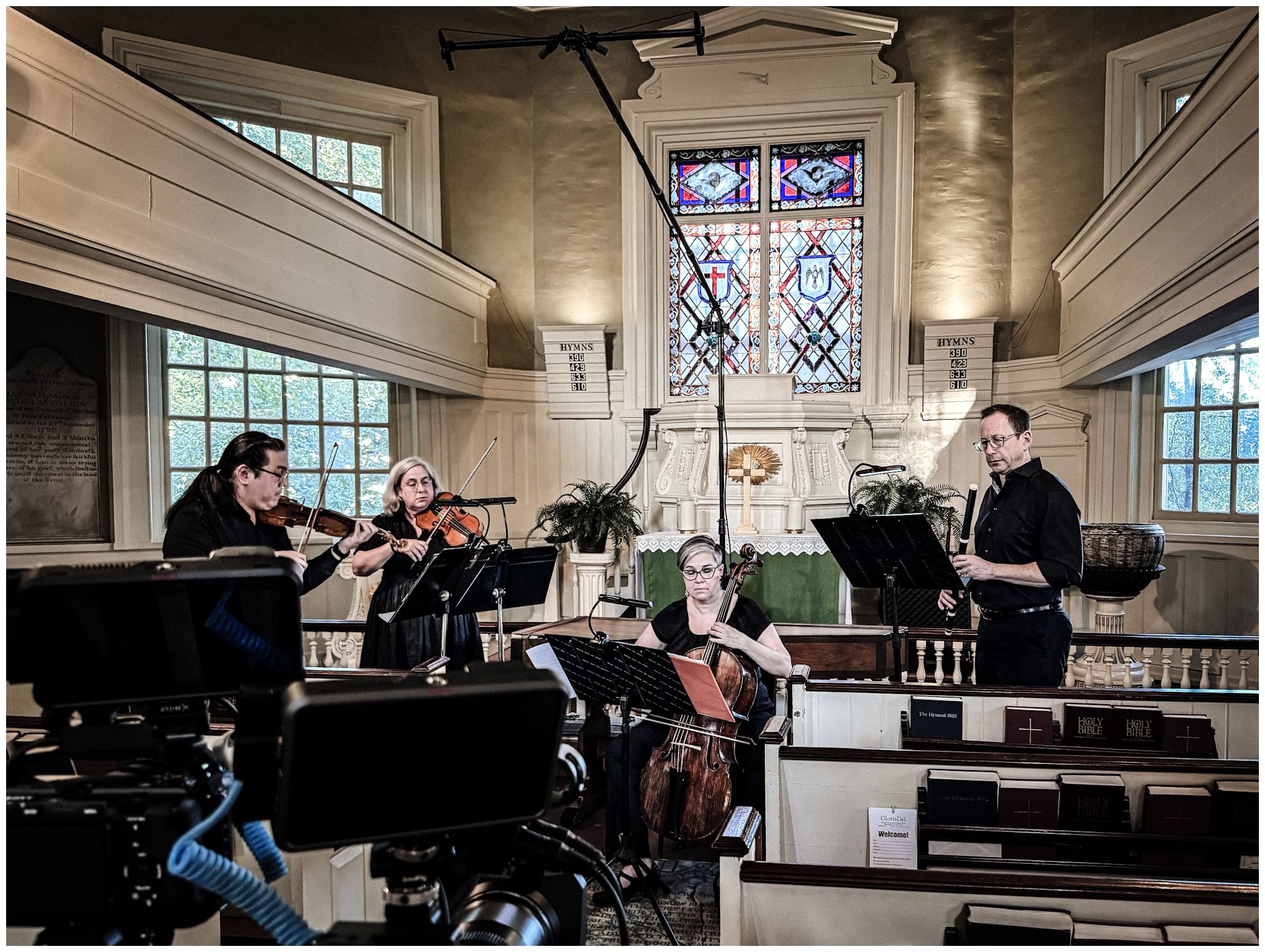 Four musicians play on the small chancel of a historic church, with a small stained glass window and lots of white molding