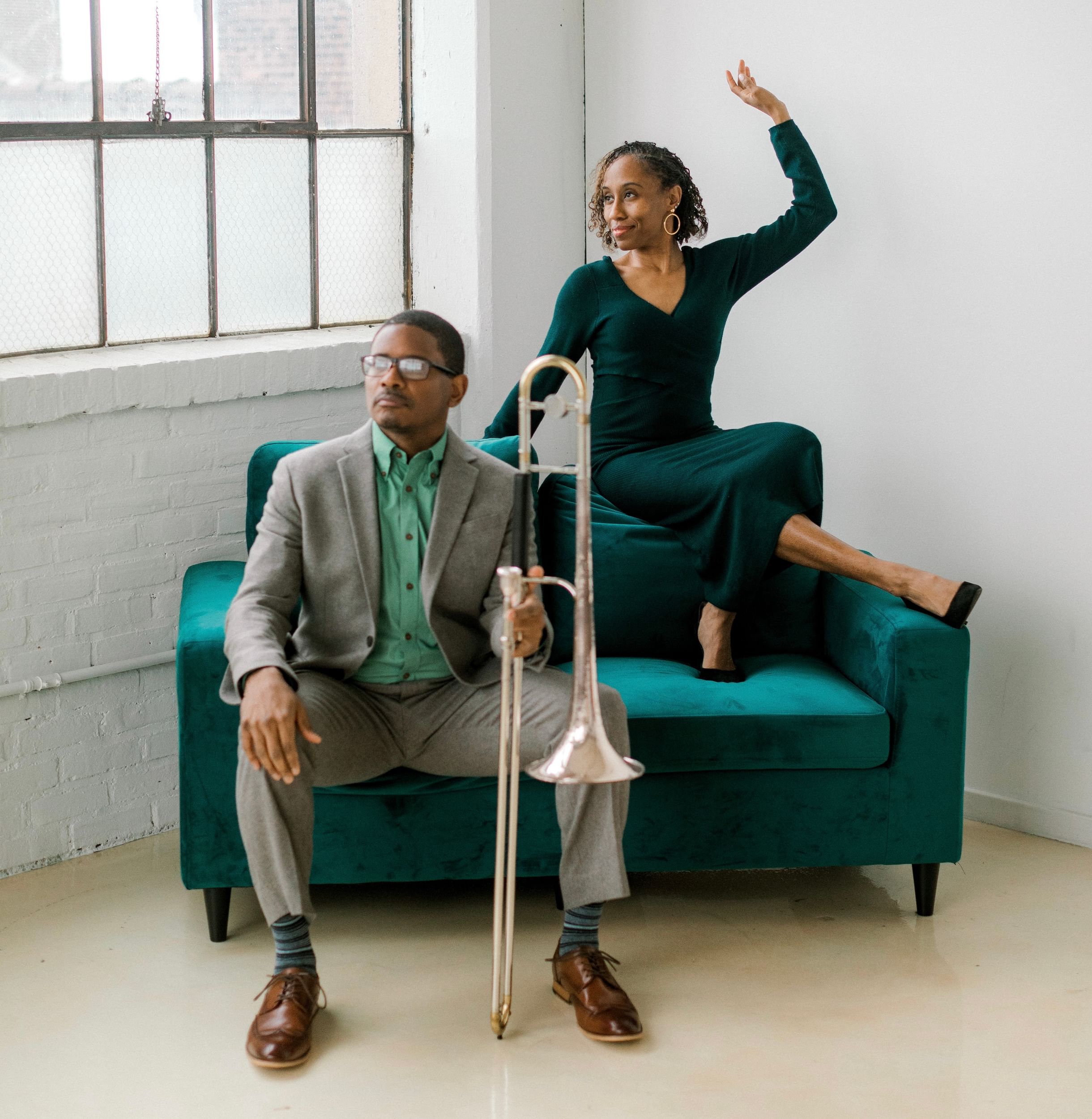 Two people on a dark blue couch. One has a trombone, the other sits on edge of chair, an arm and leg out, as if in step