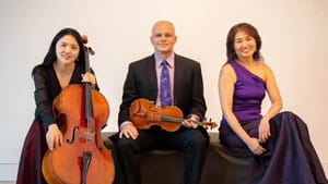 Jie Jin has a cello, Mazzocchi holds a violin, Yamazki sits with them on a bench. All are dressed in formalwear
