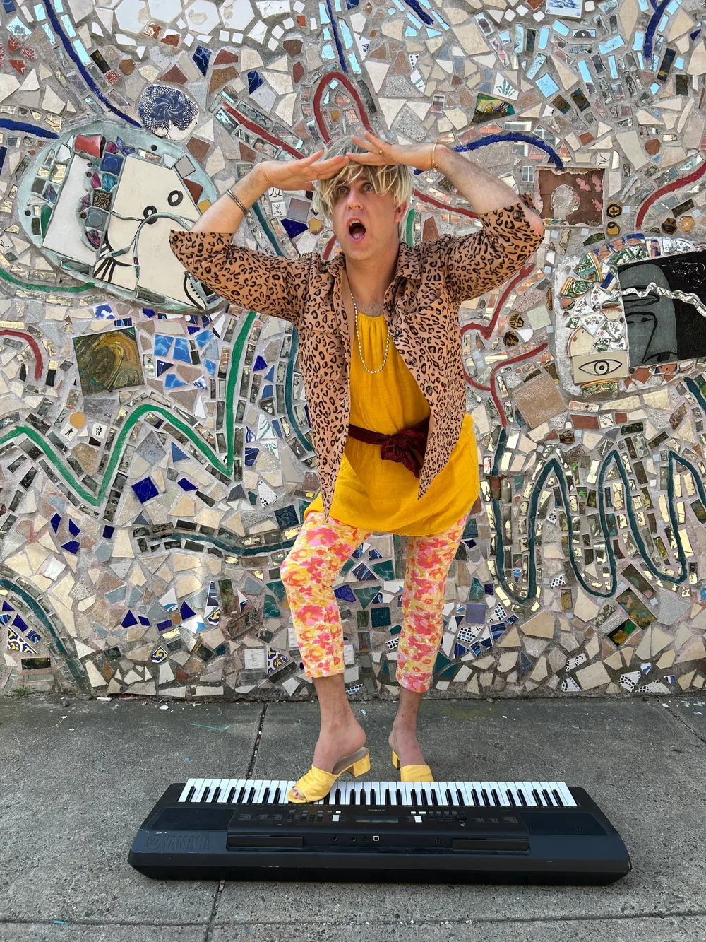 Kitrosser, in floral, yellow, leopard-print drag, poses as if he’s searching, one foot on a keyboard on the ground.