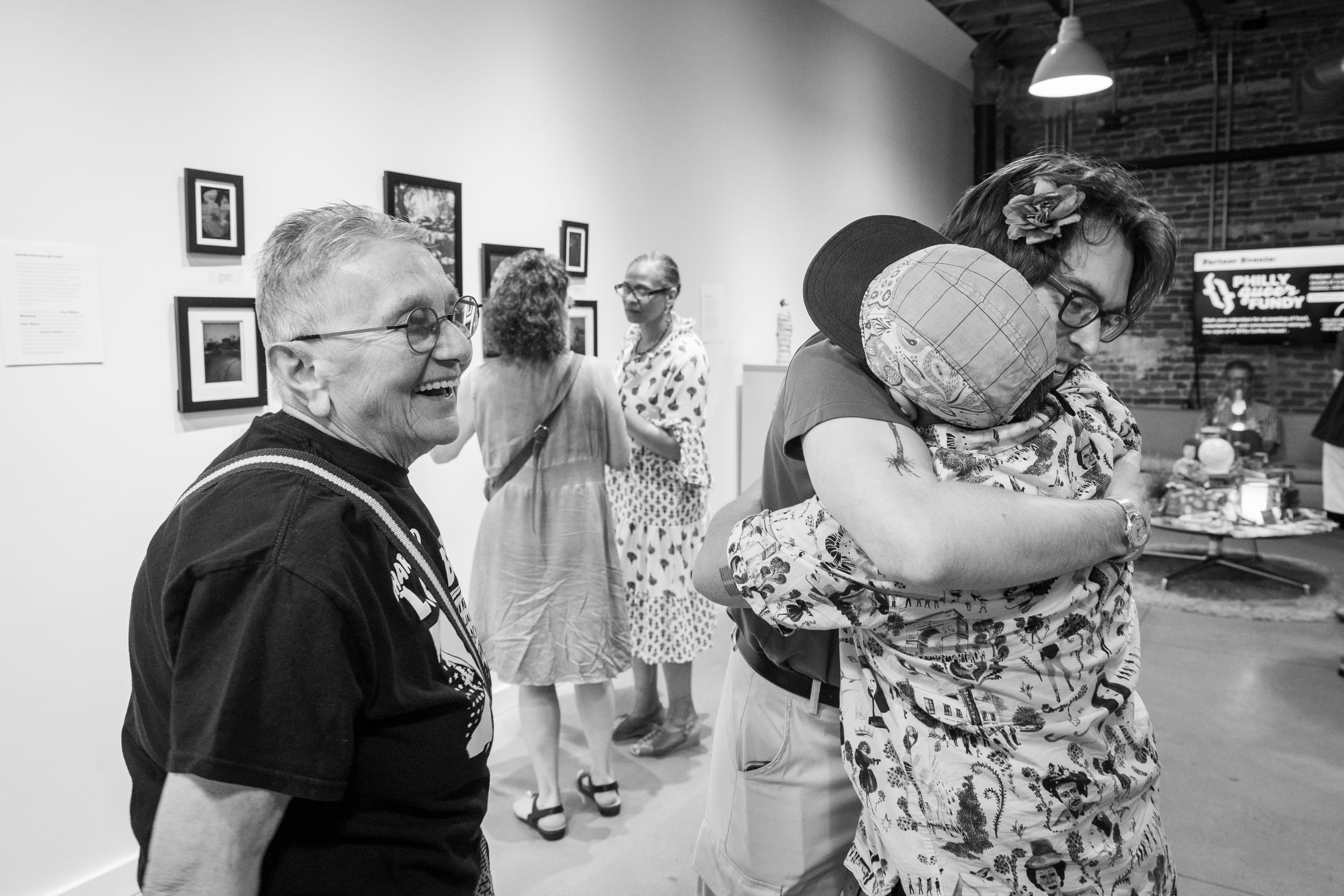 Black & white art gallery view photo with Olshan smiling in the foreground while two other people embrace.