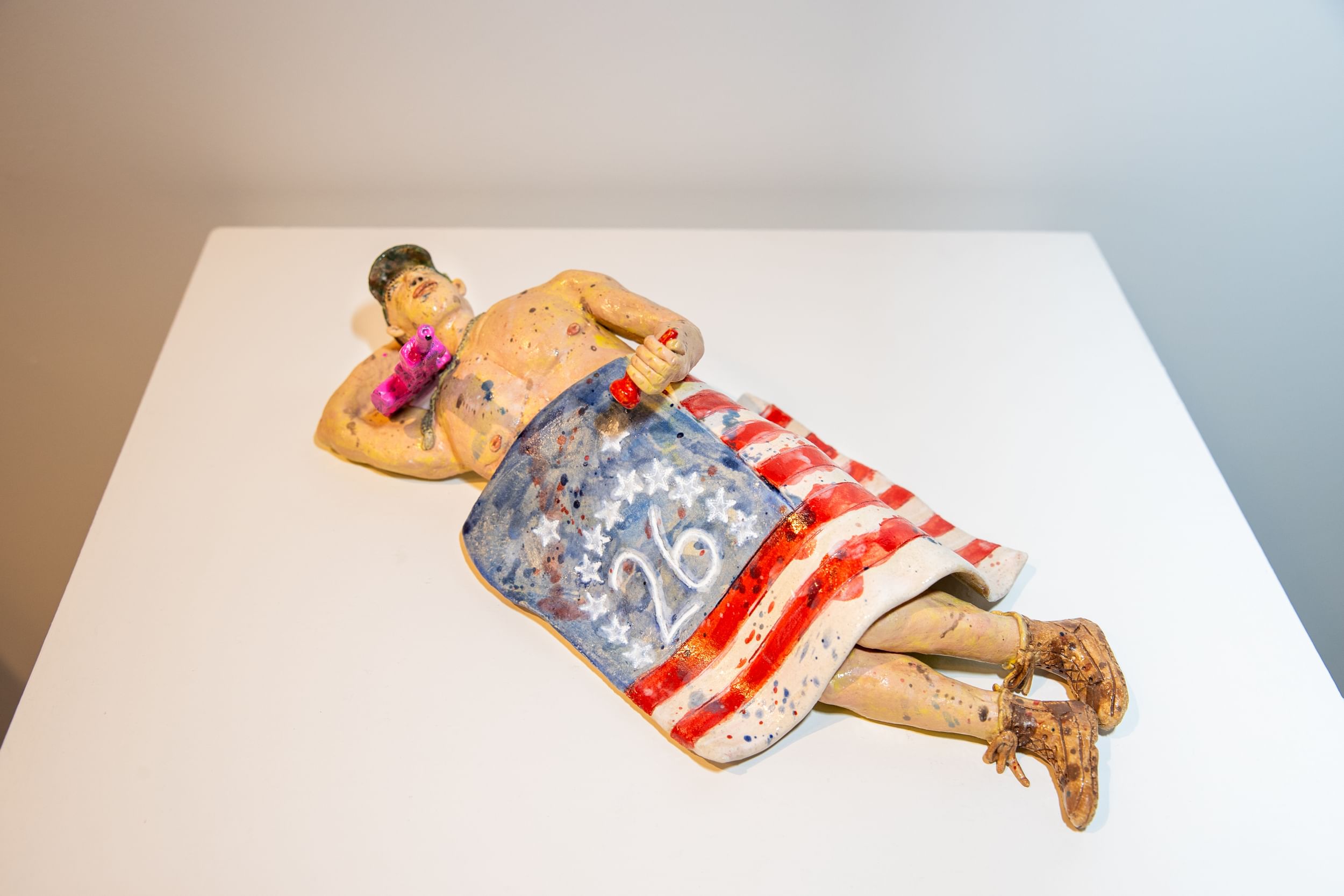 Closeup on small glazed ceramic sculpture of a person wearing only a baseball hat and boots, using a US flag as a blanket