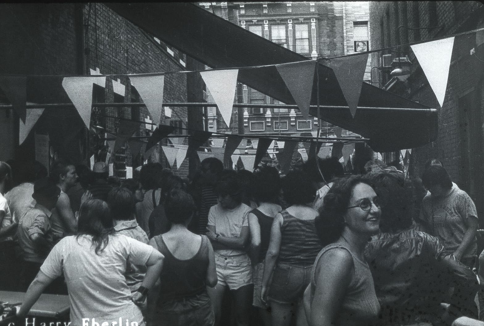 Black & white photo of a packed city alleyway hung with triangular flags over a crowd in 1970s summer clothes.