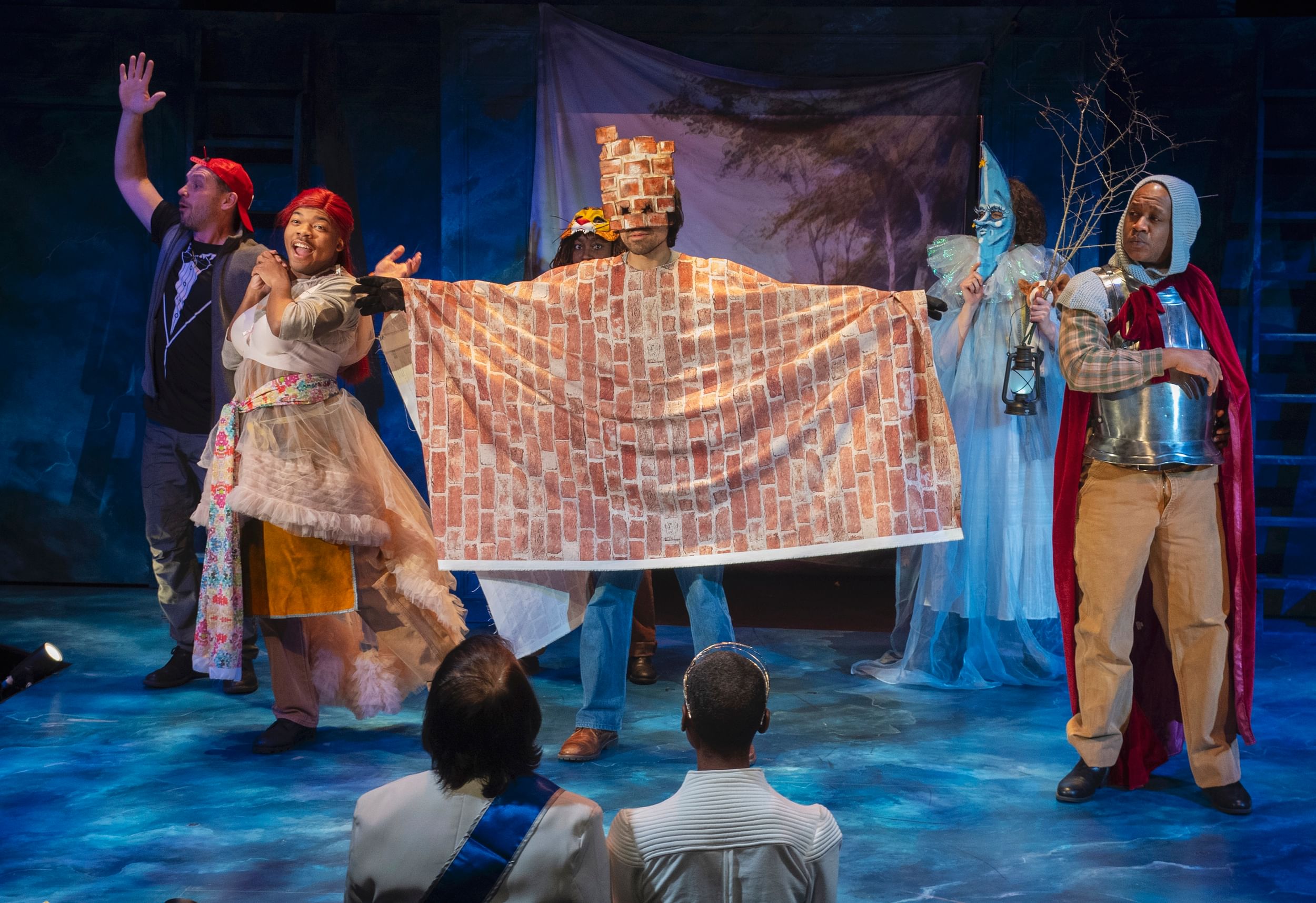 Several actors in costume on stage, one actor centered with a mask and a blanket or shawl with bricks patterned on them