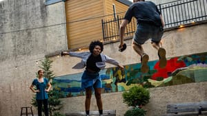 An action shot of QvK, with two actors, a Black woman and white man, leaping high in the air while swinging fencing swords.