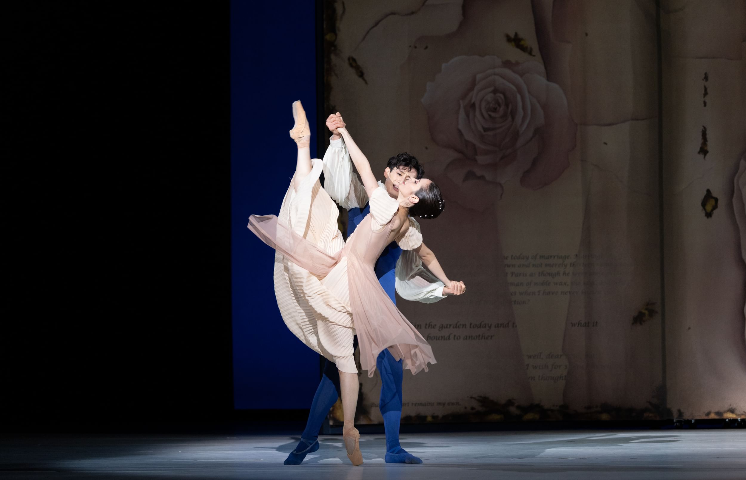 Liang, in blue behind Golz in pale pink, holds her hands as her arms extend in sweeping en point pose, one foot pointing up
