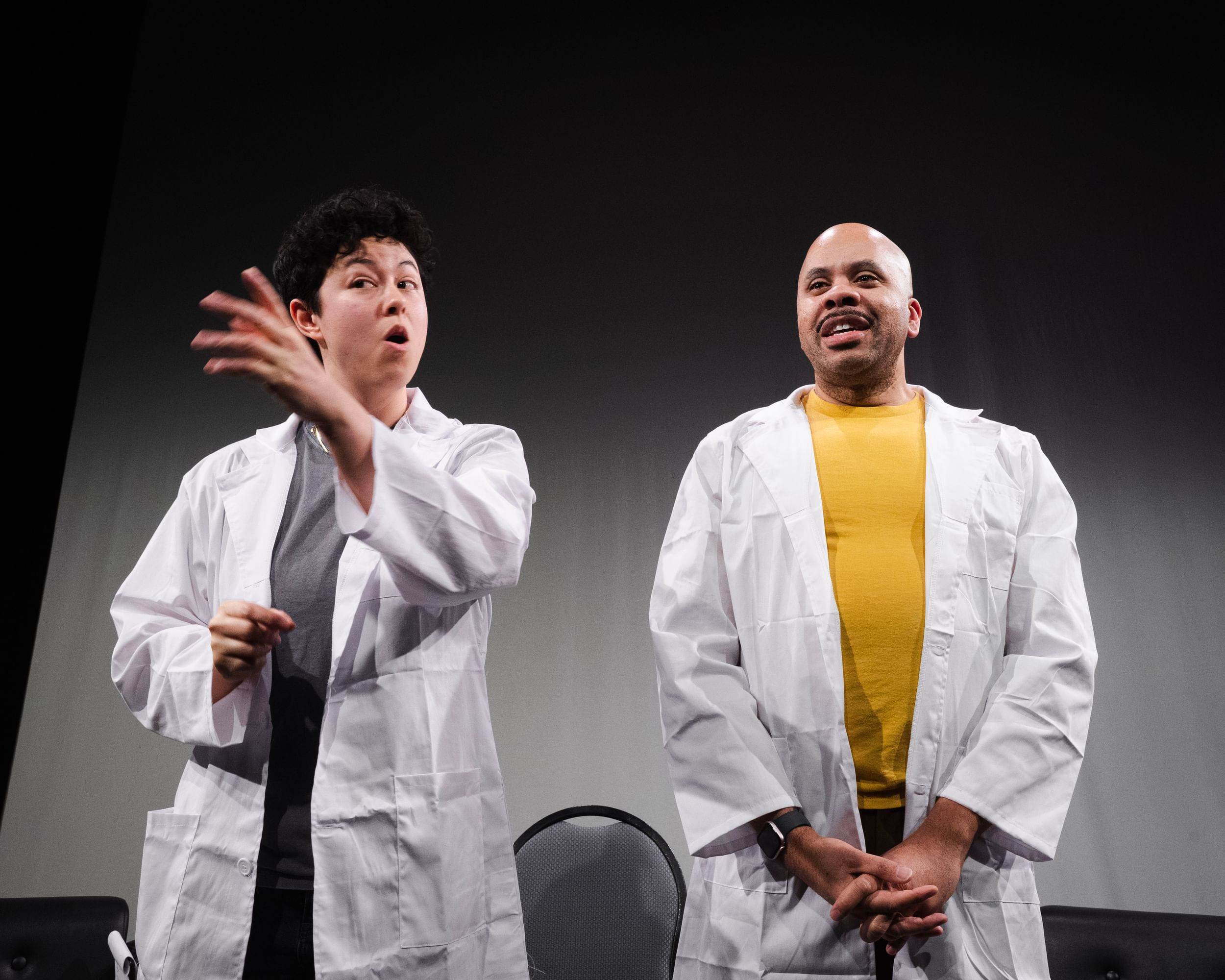 Martin and Thomas on stage next to each other, addressing the audience, both wearing white lab coats over casualwear