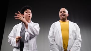 Martin and Thomas on stage next to each other, addressing the audience, both wearing white lab coats over casualwear