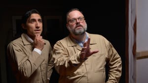 Two men in beige jumpsuits look on in wonder, eyes wide, each with one hand at their sides.