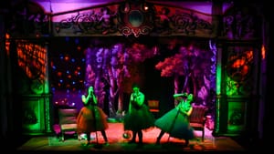 Three women in knee-length gowns on a colorfully lit stage sing into microphones like rock stars