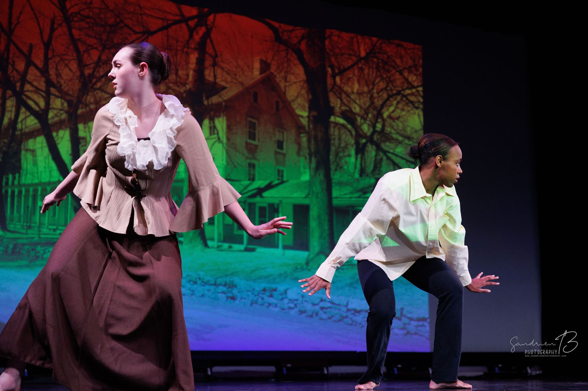A Black woman and white woman on stage, a backdrop reveals a large house shrouded in trees, both seem to be escaping