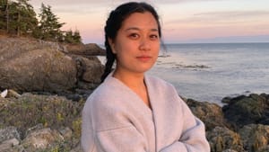 Portrait of Hsu, a Taiwanese woman. Her arms crossed in a sweater, she stands along tall, large rocks by large body of water
