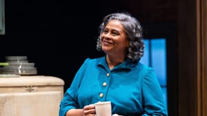 Finister, a Black woman with shoulder-length gray curls, wears a turquoise dress and smiles, holding a mug.