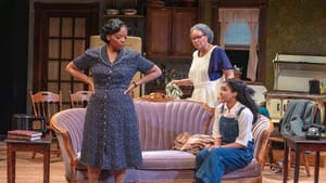 In a dotted dress, Johnson stands with hands on her hips. Wyatt sits on a couch. Fairbanks passes behind carrying a towel.