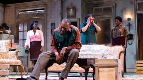 DeShields sits slumped and ashamed on a coffee table in the foreground. The women express shock & dismay in the background