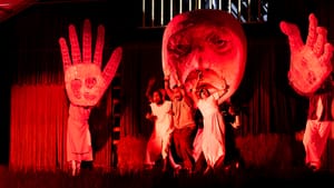 Washed with red light, puppeteers work a giant face and hands, closing in on a small group of frightened people at center.