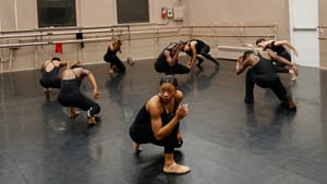 Joseph in all black tights crouches in a rehearsal space, turning away from the camera. Other dancers look away, in mid-pose