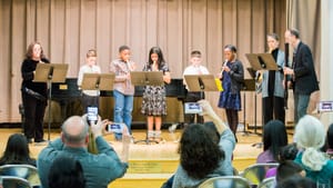 Six kids and two adults at podiums play recorders on stage. Audience members hold up their phones, taking pictures