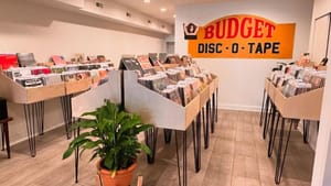 The browsing area, with wooden record displays in a bright white space and a vintage “budget disc o tape” sign on the wall