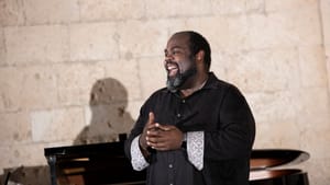 Mobley, a Black man, smiles while standing in front of piano, wearing all black top with white and gray sleeves