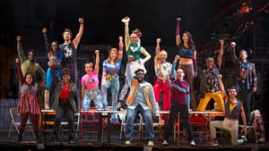 A scene from Rent: 15 ensemble members in colorful 90s garb standing on an around a long table, raising one fist in the air.