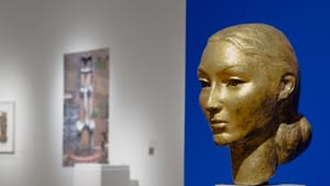 Graceful gold-toned metal bust of a beautiful woman’s face, against a blue backdrop in the gallery.