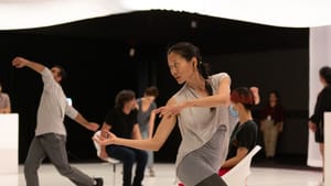 In the foreground, Chang, in gray, moves gracefully with arms extended; ensemble and audience members sit and move behind her