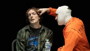Scammell, in a head bandage and orange jumpsuit, menaces McLenigan, in a plaid shirt, holding knuckles to his head like a gun