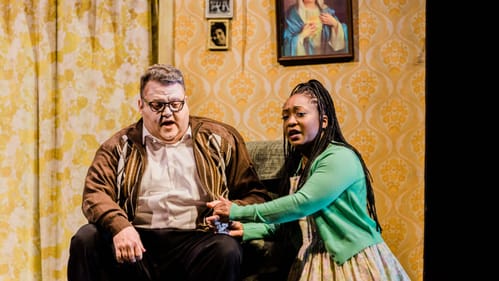 Scene from opera. Evans, a white man in a brown jacket, in an armchair next to McMillon, a Black woman, in a green sweater.