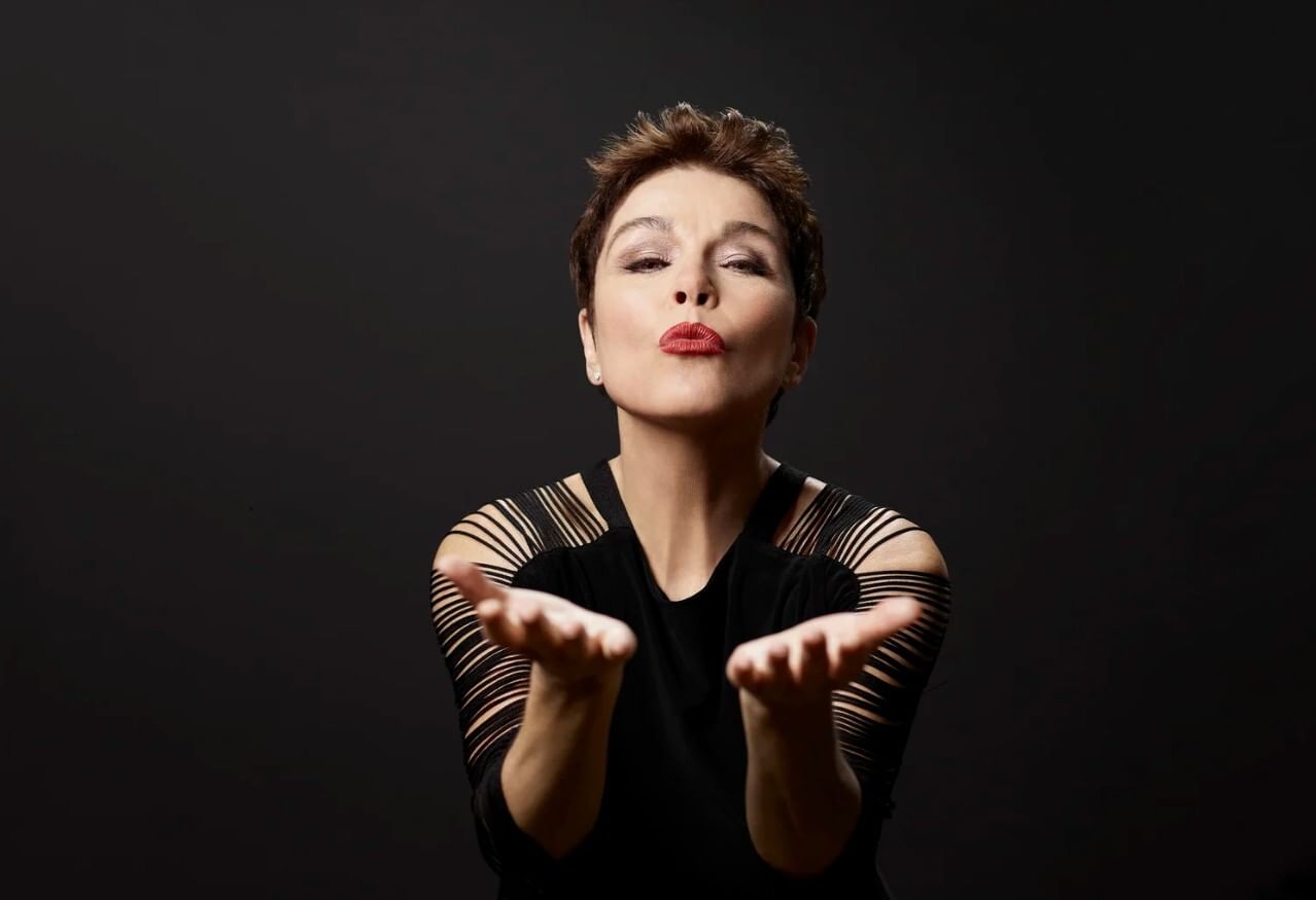 Andreas, a white woman with short brown hair, dramatically blows a kiss at the viewer.