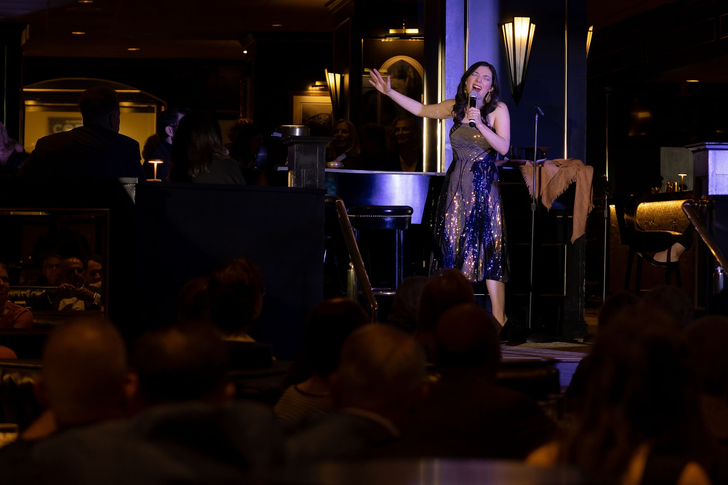 Benko, a white woman in a sleeveless sequined evening gown, sings passionately into a mic at a ritzy restaurant cabaret.
