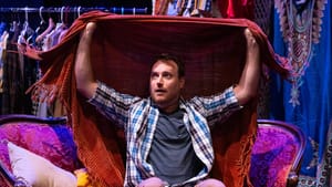 Droxler, a white man in a plaid button-down, sits on a couch looking slightly afraid, holding a fringed blanket above him