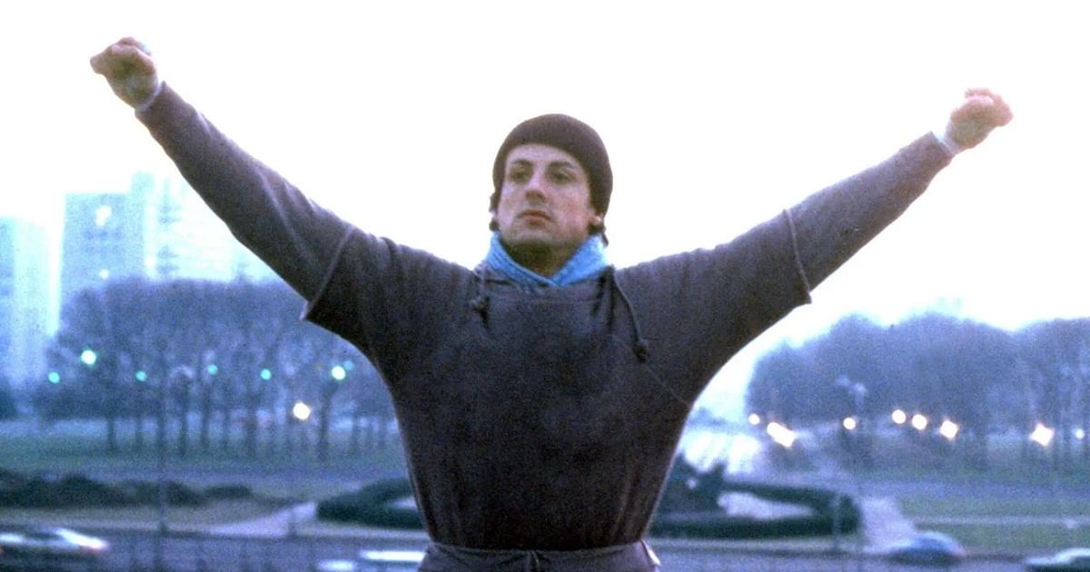 A white man in a gray sweater and beanie holds his hands up on the steps of the Philadelphia Museum of Art.