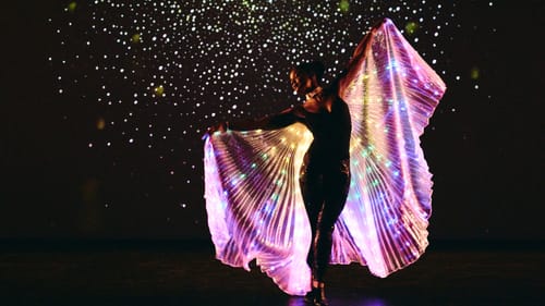 Coats, a Black woman, poses on tip toe with a colorfully lit iridescent pleated fabric held up like wings with stars behind