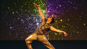 Lee, a Black man, in gold pants & patterned gold tank, poses with arms gracefully outspread with a colorful starry backdrop