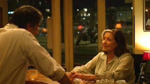 Movie still. A woman, mid-age or older, looks on at a man at what looks like a bar area, windows showing the street behind