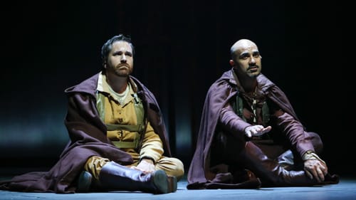 The two actors sit cross-legged on the stage, looking defeated, both wearing purple cloaks over Elizabethan garb..
