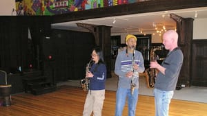 Three people playing wind instruments stand next to each other in an indoor area