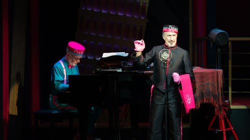 Close up on Gilbert playing the piano next to Ernst standing at a vintage mic. They wear round hats and satiny ship livery.