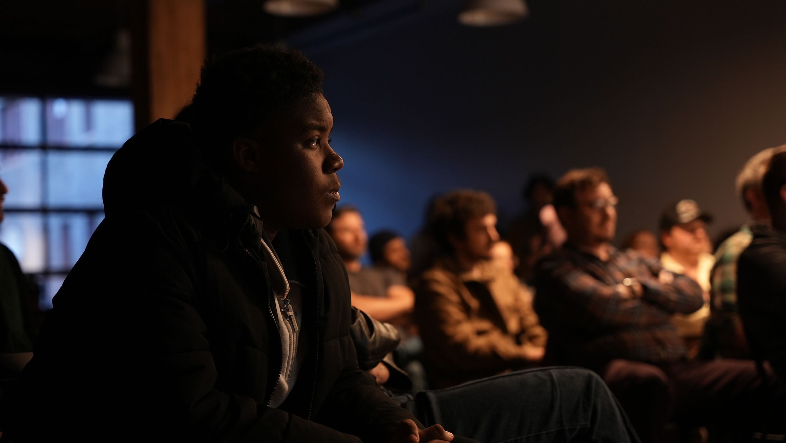 Closeup on a Black audience member in profile looking thoughtfully ahead, with many other attendees in the background.