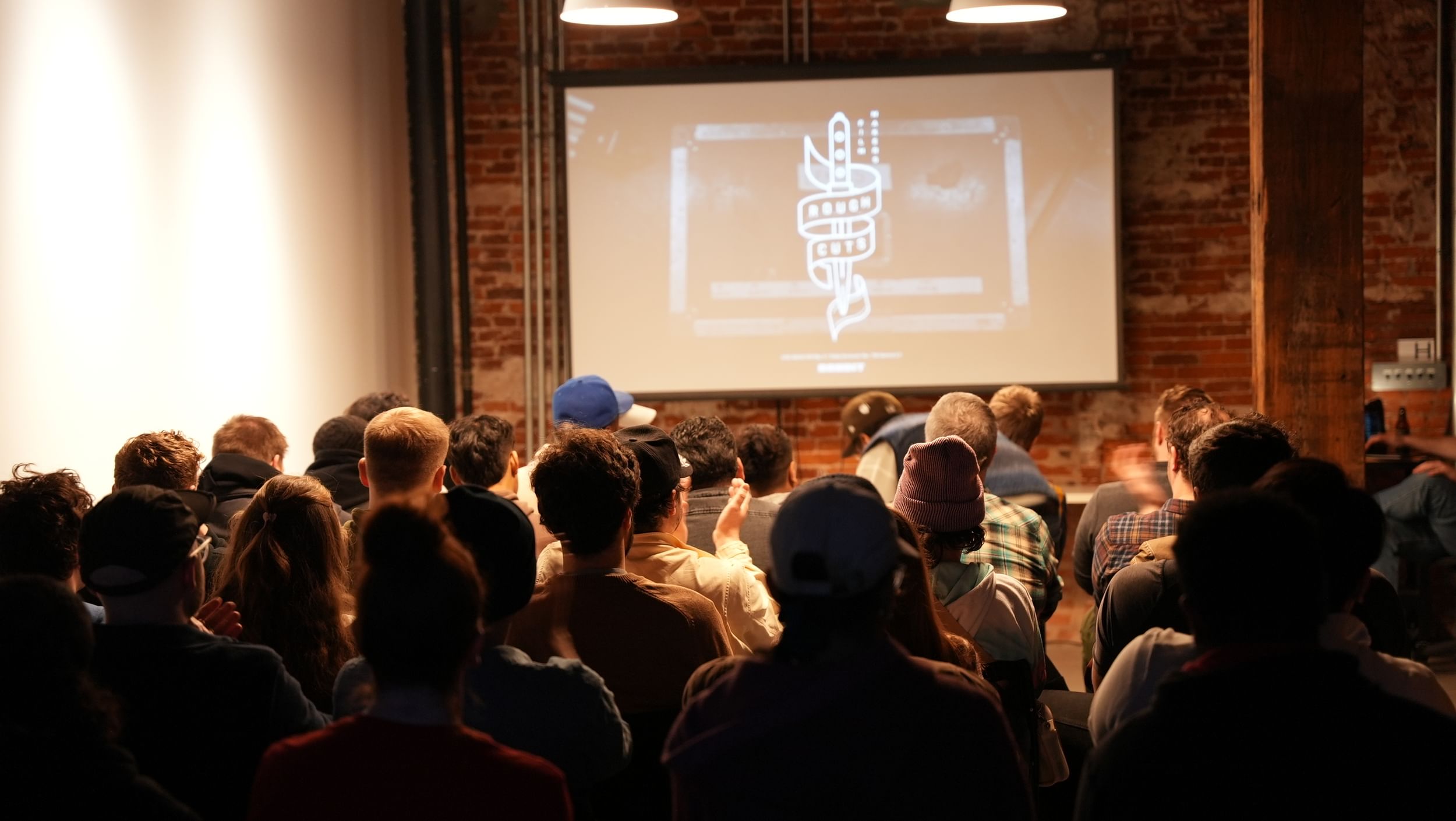 View from behind of a few dozen people seated in a brick-walled gallery, facing a projection screen.