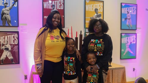 A Black family of 4 in Kwanzaa shirts smile together in front of a wall of Rucker portraits in the style of Wheaties boxes.