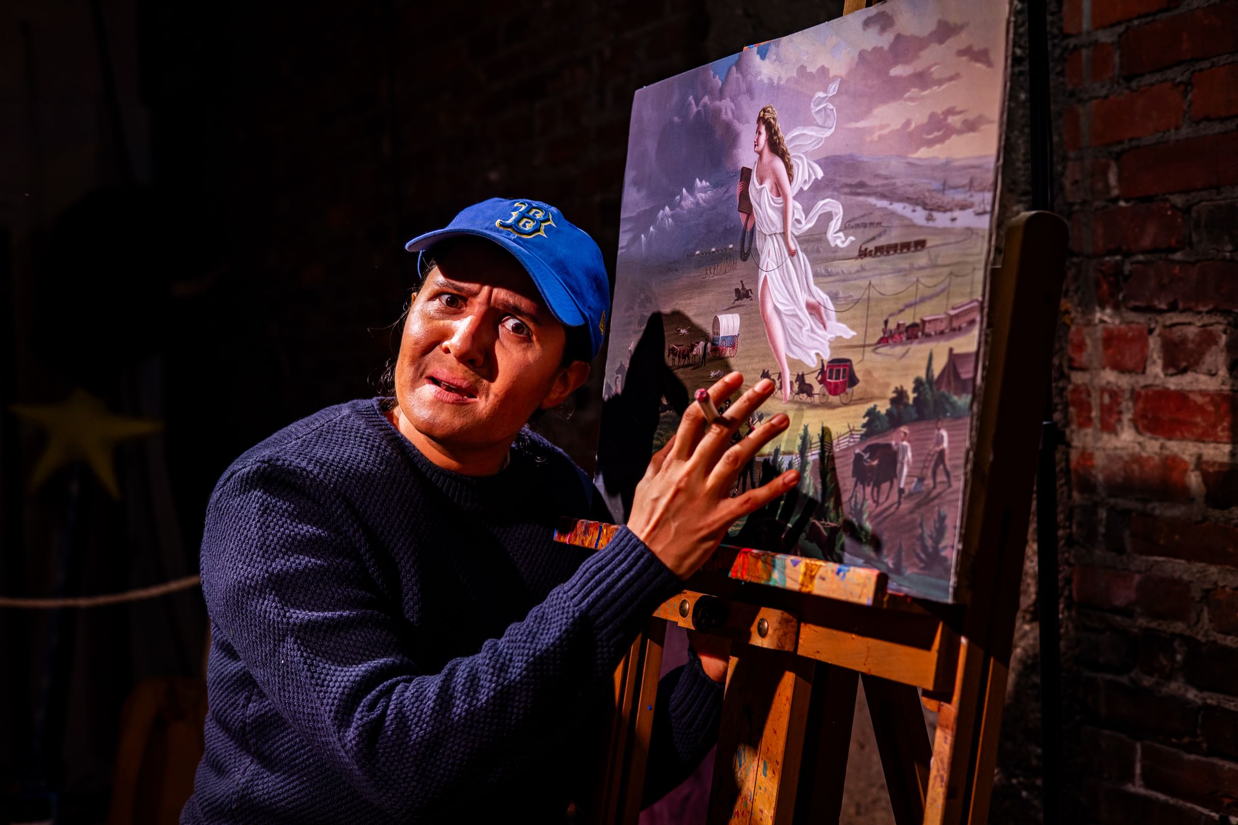 Sazak, in a Boston ballcap & blue sweater, holds a cigarette and gestures fervently to a reproduction of ‘American Progress’