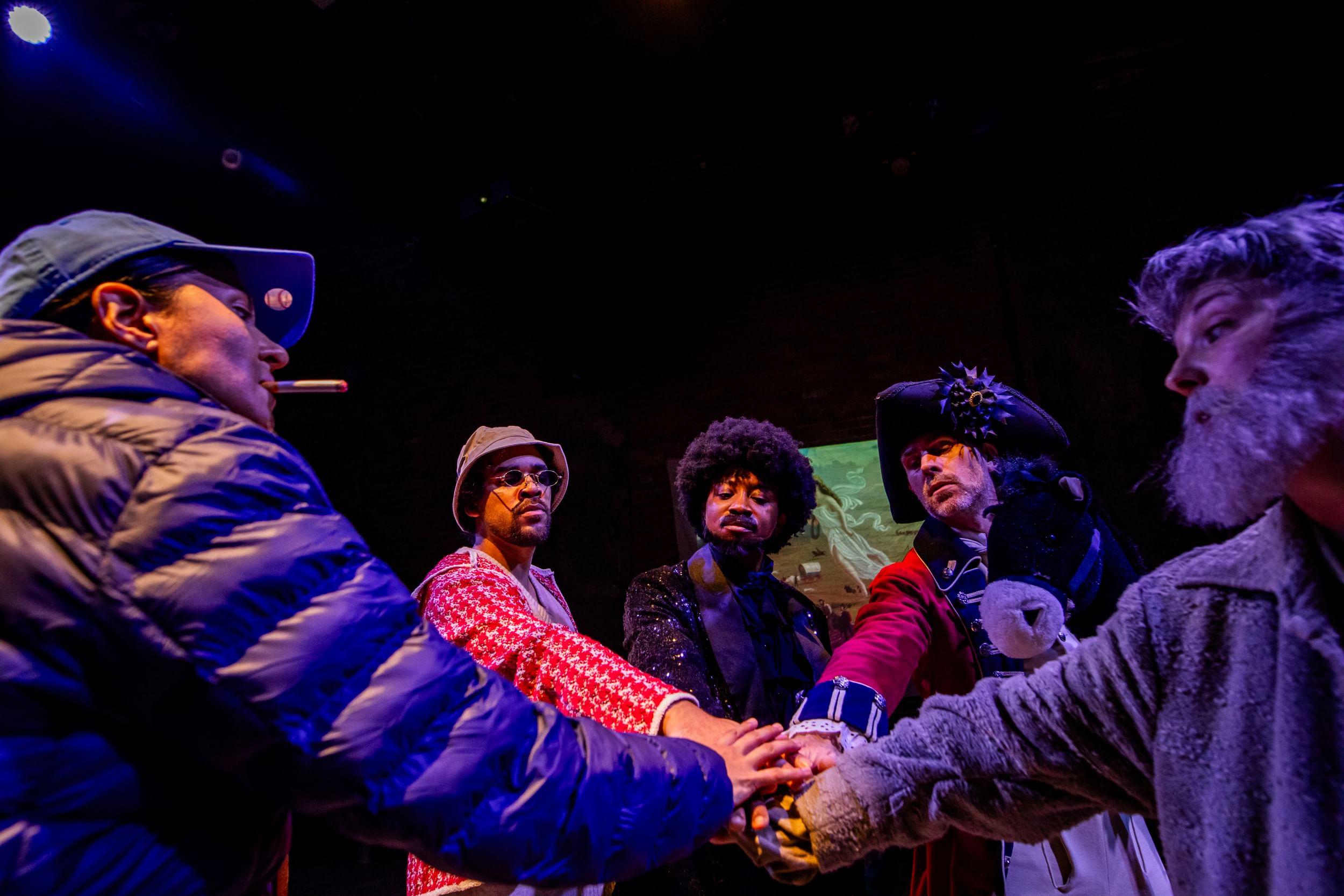 Five diverse actors, in various garments by era, pose seriously like a wheel with their hands meeting in the center.