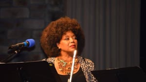 Floyd, a Black woman, with an afro, at a podium with a flute.