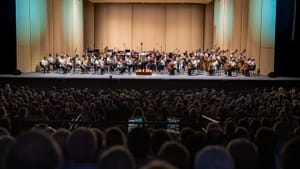 Full orchestra onstage at SPAC, all in white shirts. They appear tiny, in front of hundreds of silhouetted audience members.