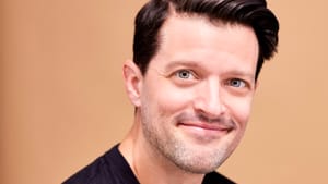 A headshot of Pacek, in a black shirt smiling against a soft pink colored backdrop
